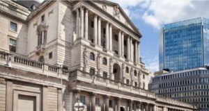 Photograph of the historic Bank of England building in London, with double-decker buses passing by — symbolizing the central bank’s role in shaping UK financial policy and the future of the digital pound.
