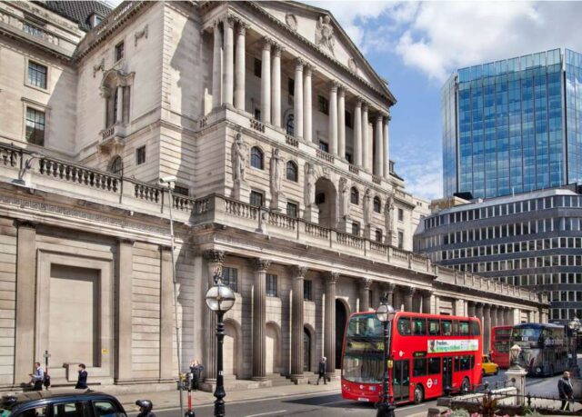 Photograph of the historic Bank of England building in London, with double-decker buses passing by — symbolizing the central bank’s role in shaping UK financial policy and the future of the digital pound.